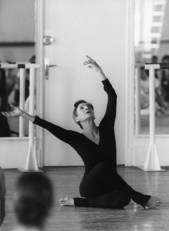 bella in a black leo and tights sitting one leg crossed over the other with arms above her head, and her face tilted upward. she is teaching a dance class, leading in front of the mirror.